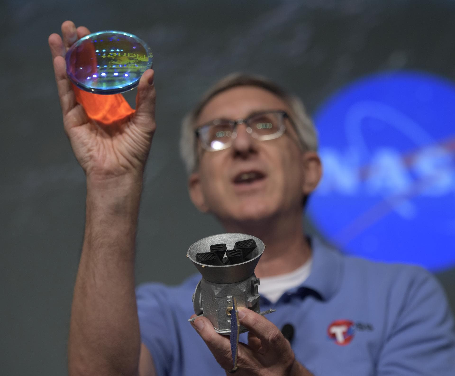 Jeff Volosin, TESS project manager, NASA’s Goddard Space Flight Center, holds a spare camera lens and a model of the Transiting Exoplanet Survey Satellite (TESS) during a media briefing, Wednesday, Ma