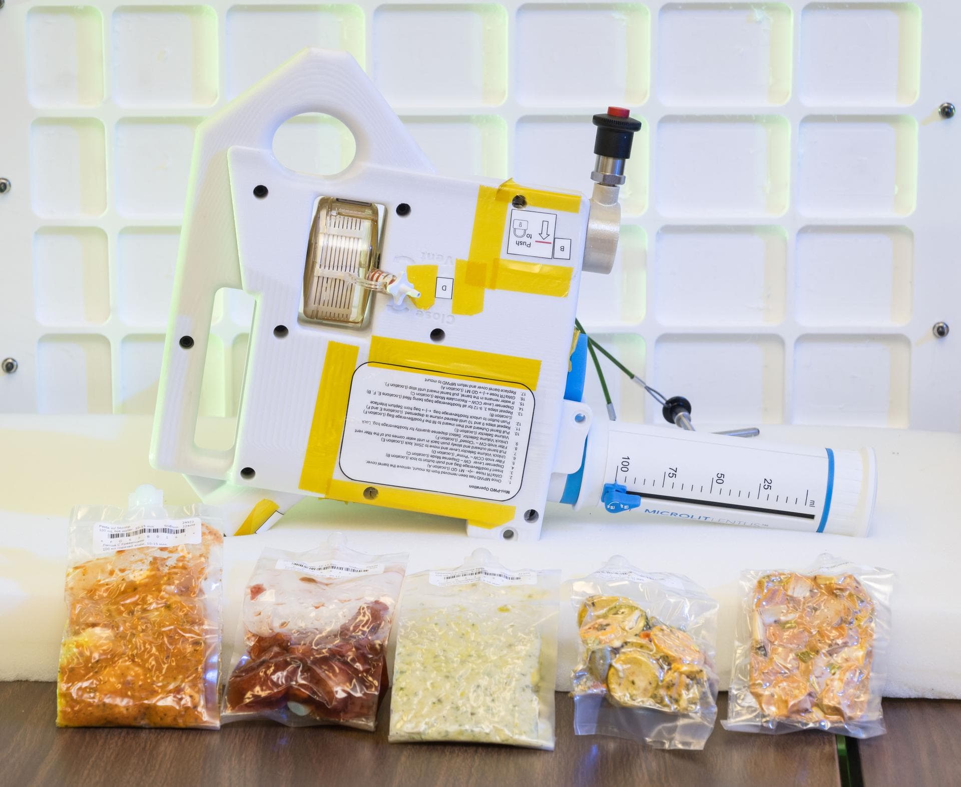 jsc2024e040685 -- A prototype of the Mini Potable Water Dispenser, currently in development at NASA’s Marshall Space Flight Center, is displayed alongside various food pouches during a demonstration a