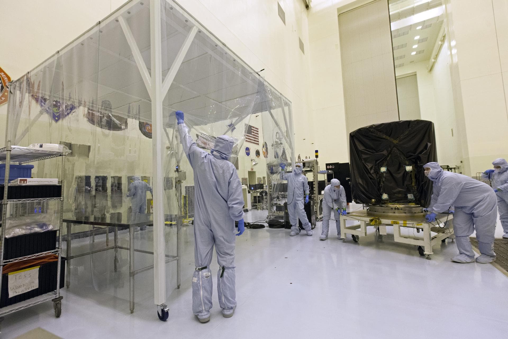 Technician dressed in clean room suits move NASA's Transiting Exoplanet Survey Satellite (TESS) on a test stand to a clean room tent inside the Payload Hazardous Servicing Facility (PHSF) at the agenc