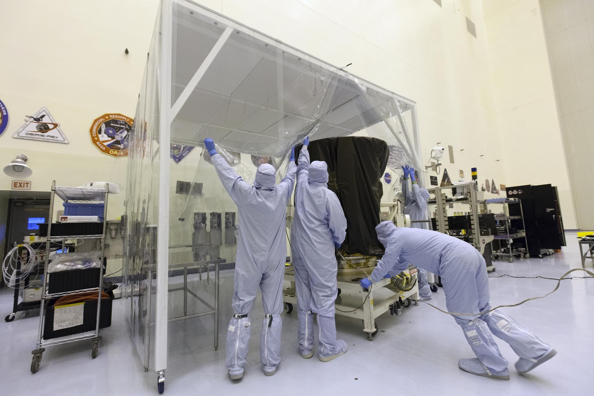 Technician dressed in clean room suits move NASA's Transiting Exoplanet Survey Satellite (TESS) on a test stand into a clean room tent inside the Payload Hazardous Servicing Facility (PHSF) at the age