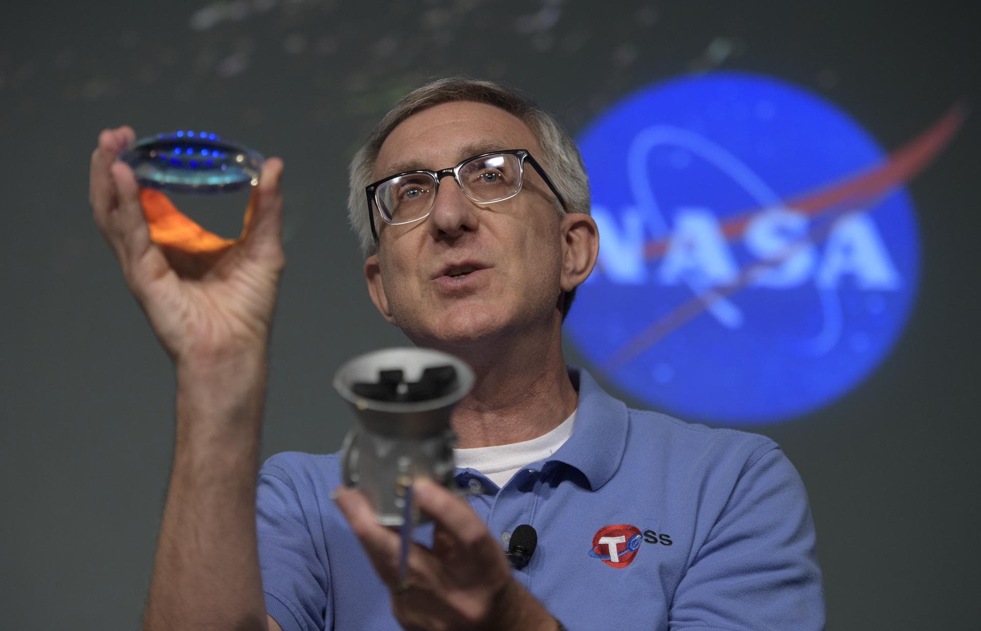 Jeff Volosin, TESS project manager, NASA’s Goddard Space Flight Center, holds a spare camera lens and a model of the Transiting Exoplanet Survey Satellite (TESS) during a media briefing, Wednesday, Ma